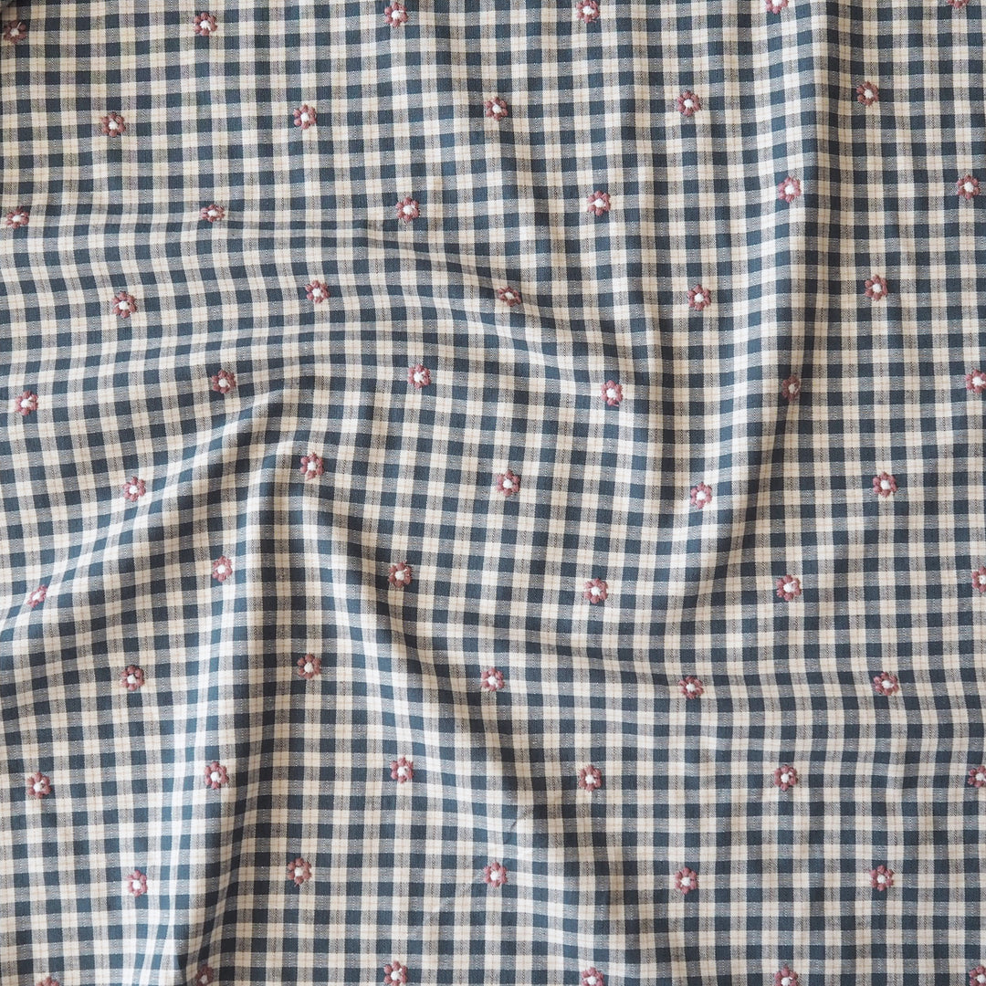 A close-up of a fabric surface with a small gingham check pattern in navy and off-white, overlaid with a scattered motif of tiny pink and white flowers.