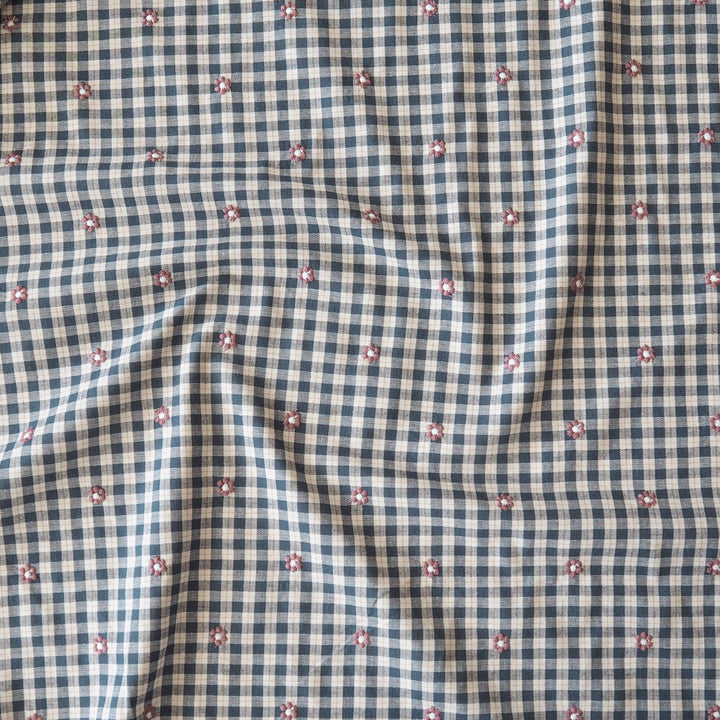 A close-up of a fabric surface with a small gingham check pattern in navy and off-white, overlaid with a scattered motif of tiny pink and white flowers.