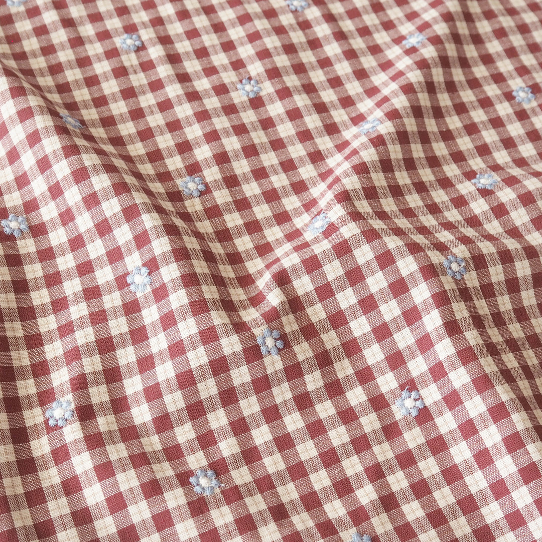 A close-up of a red and beige gingham fabric with small light blue embroidered floral motifs scattered throughout.