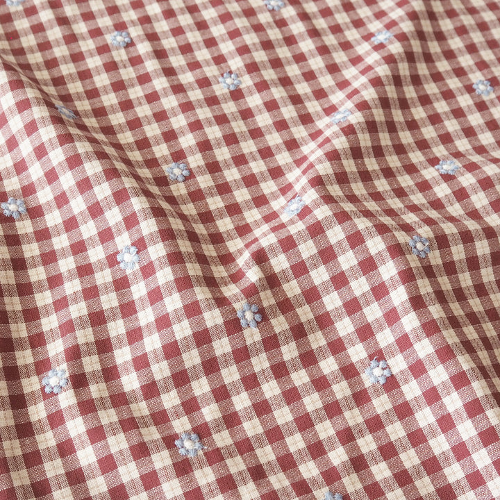 A close-up of a red and beige gingham fabric with small light blue embroidered floral motifs scattered throughout.
