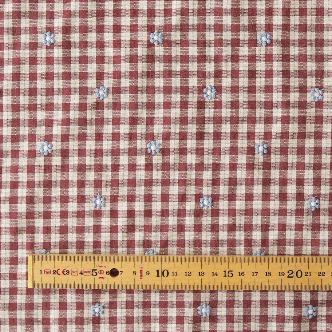 A close-up of a red and beige gingham fabric with small blue and white floral motifs, shown with a yellow measuring tape at the bottom for scale.