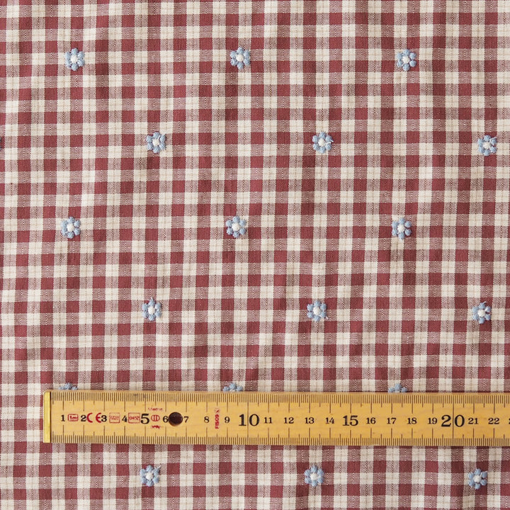 A close-up of a red and beige gingham fabric with small blue and white floral motifs, shown with a yellow measuring tape at the bottom for scale.