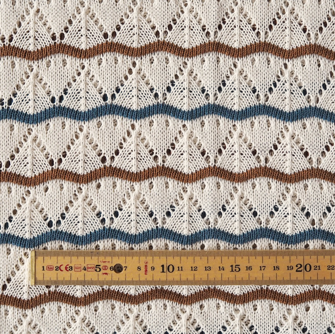 Close-up of a cream-colored knit fabric with a textured openwork pattern, featuring horizontal wavy stripes in brown and blue, with a wooden ruler at the bottom for scale.