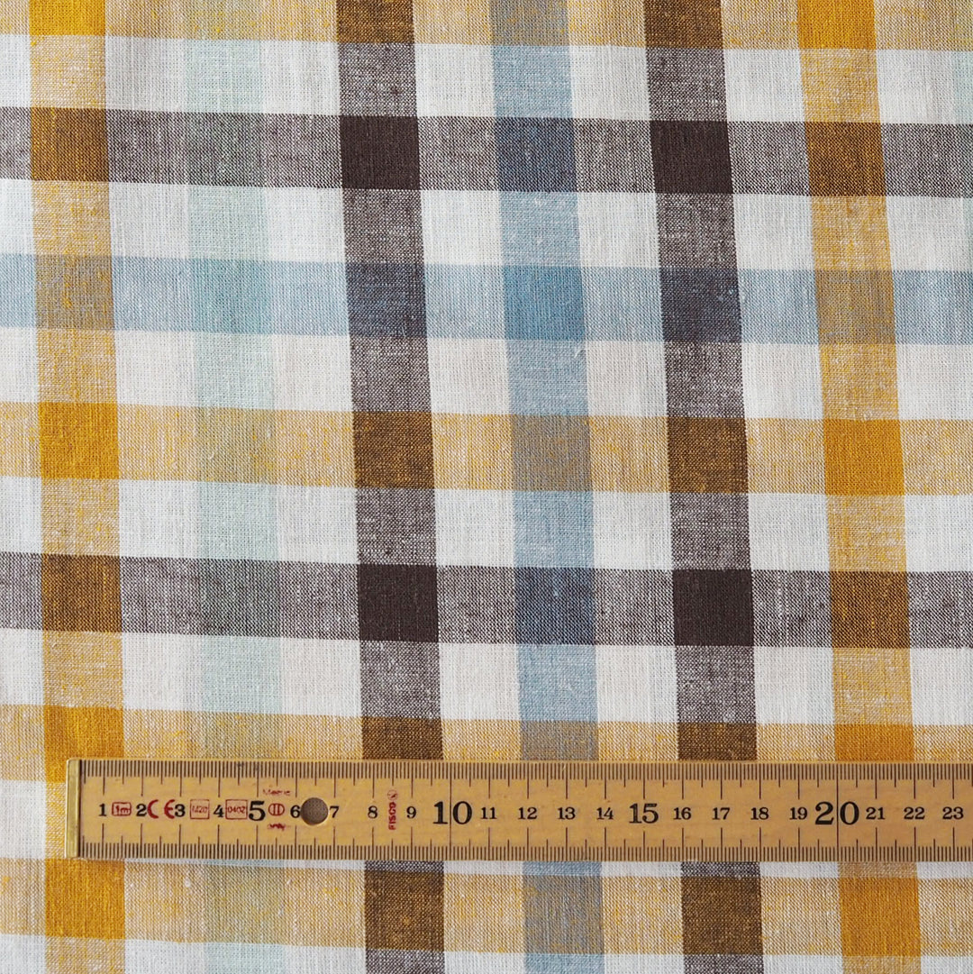 A close-up of a plaid fabric pattern with yellow, brown, light blue, and white stripes, shown with a wooden ruler along the bottom edge for scale.