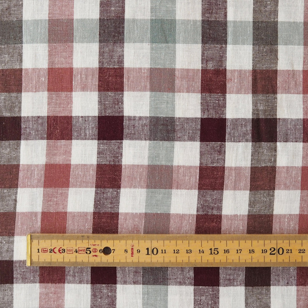 A close-up of a plaid fabric pattern with intersecting vertical and horizontal stripes in muted red, dusty rose, sage green, dark burgundy, and off-white. A wooden ruler is placed along the bottom edge to show scale.