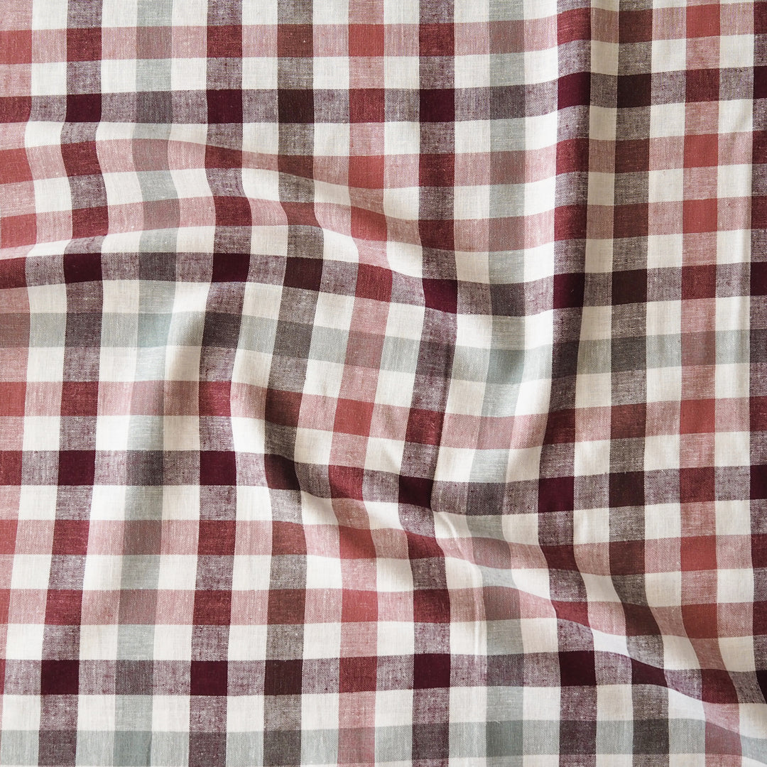 A close-up of a plaid fabric with a gingham pattern in dusty red, dark burgundy, light gray, and off-white, shown with soft folds.