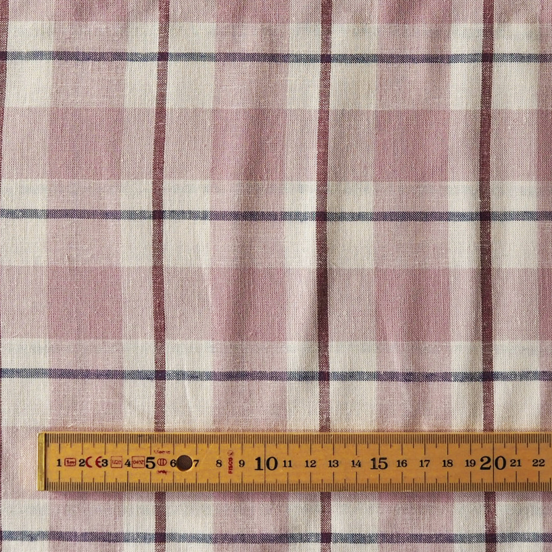 A close-up of a plaid fabric with pink, beige, and dark blue stripes, with a wooden ruler at the bottom showing the scale of the pattern.
