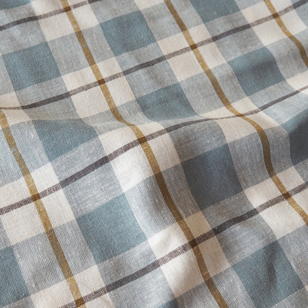 A close-up of a plaid fabric with a dusty blue, cream, and brown pattern, showing the woven texture and soft folds.