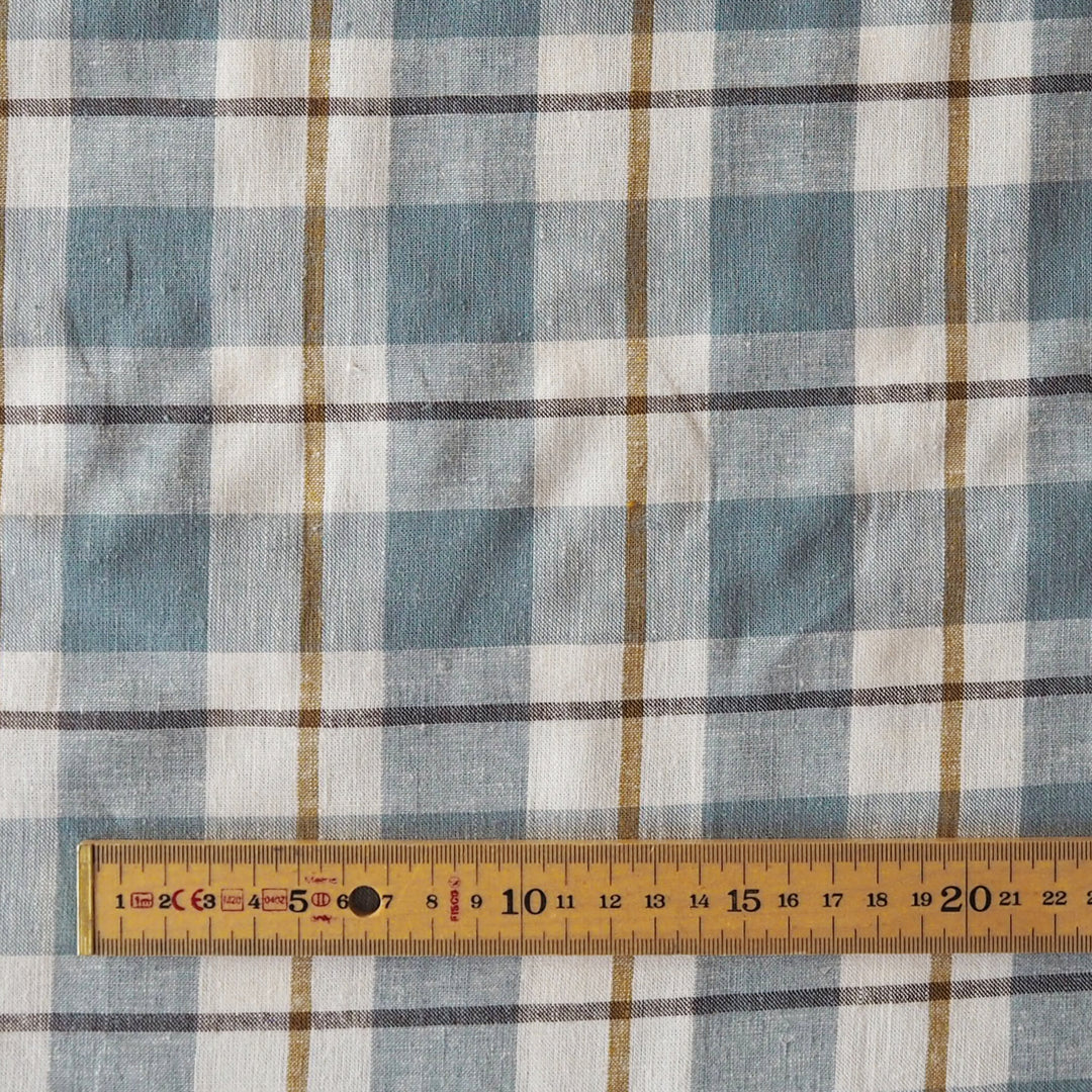 A close-up of a plaid fabric pattern in muted blue, white, and brown, with a wooden measuring tape ruler laid horizontally along the bottom edge to show scale.