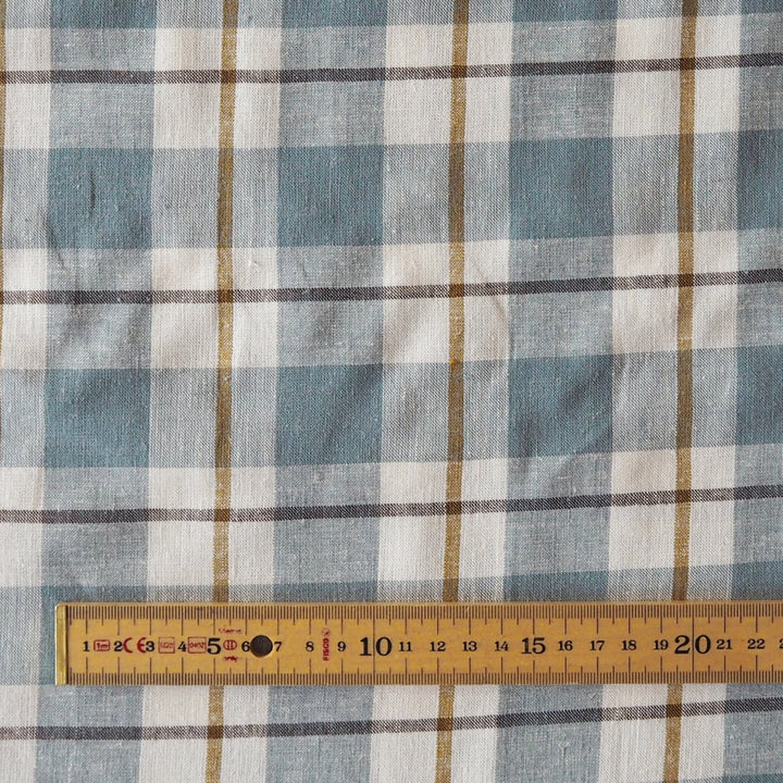 A close-up of a plaid fabric pattern in muted blue, white, and brown, with a wooden measuring tape ruler laid horizontally along the bottom edge to show scale.