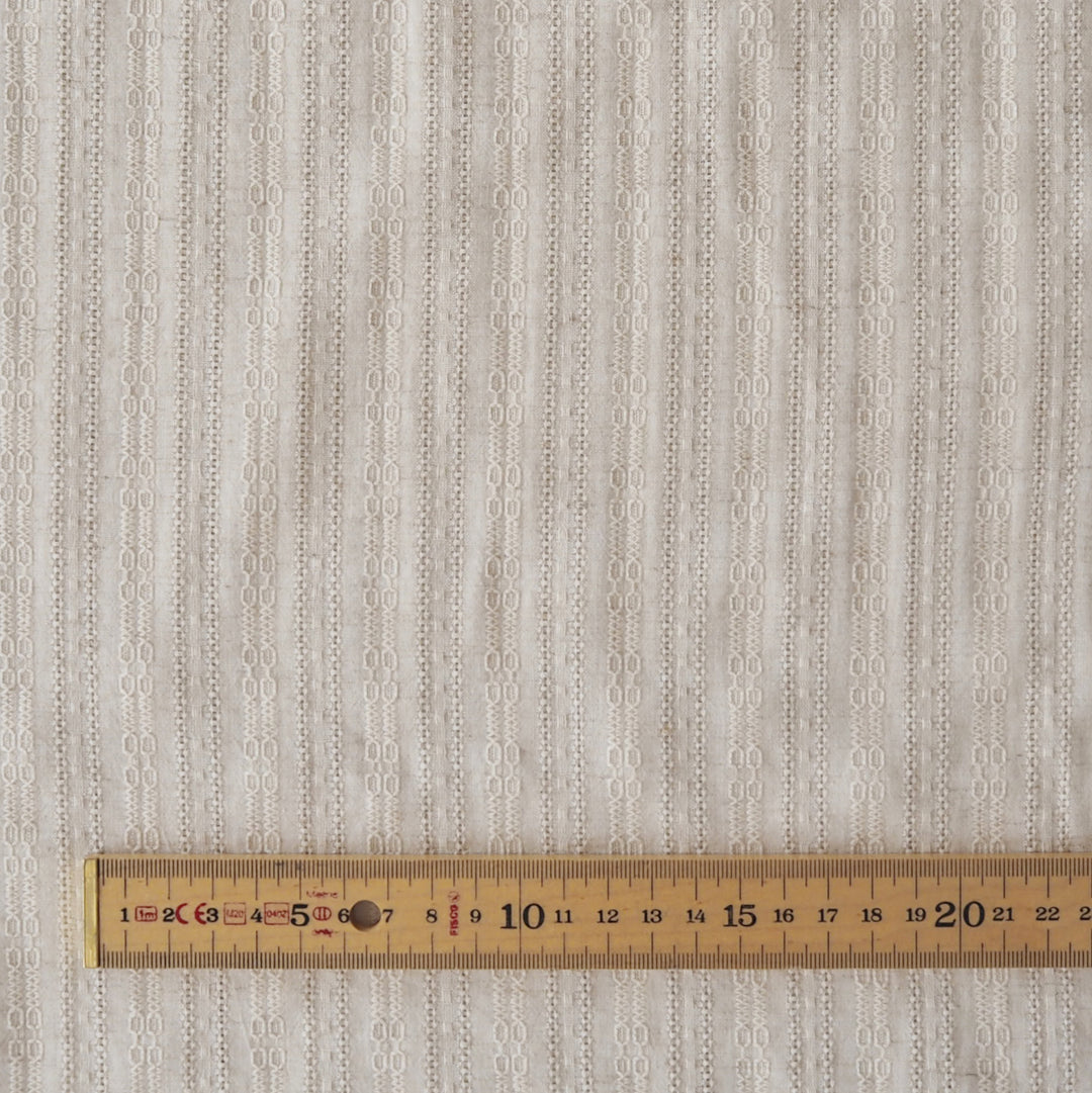 A close-up of a textured fabric with a vertical striped pattern of interlocking oval shapes in light beige and off-white, with a wooden ruler at the bottom for scale.