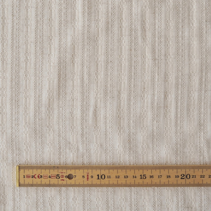 A close-up of a textured fabric with a vertical striped pattern of interlocking oval shapes in light beige and off-white, with a wooden ruler at the bottom for scale.