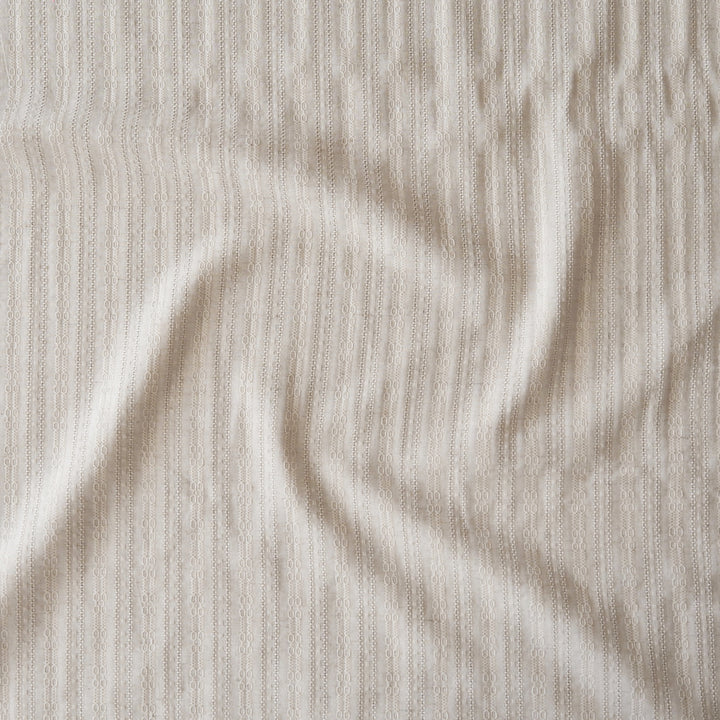 A close-up of a textured, ribbed fabric in a warm off-white or cream color, shown with soft folds and shadows.