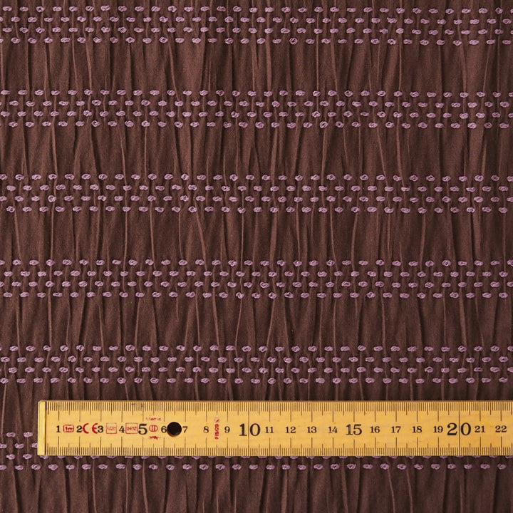 Brown pleated fabric with horizontal rows of small pink embroidered dots, shown with a yellow measuring ruler at the bottom to indicate scale.