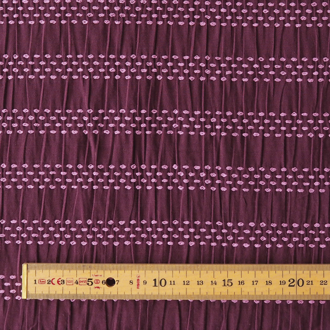 A close-up of a dark purple fabric with horizontal rows of small light pink embroidered dots, shown with a wooden ruler at the bottom for scale.