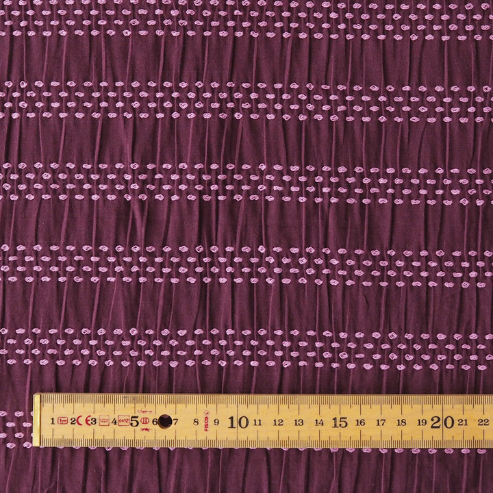 A close-up of a dark purple fabric with horizontal rows of small light pink embroidered dots, shown with a wooden ruler at the bottom for scale.