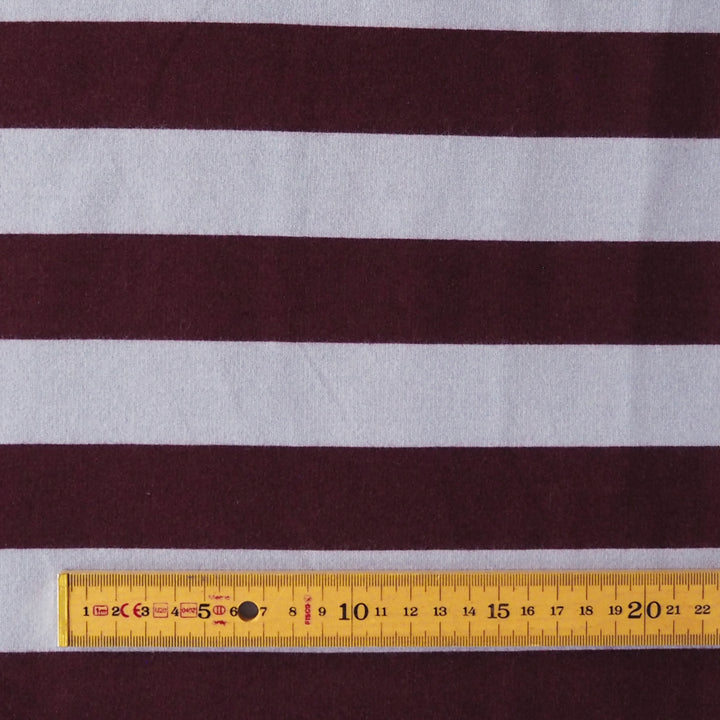 A close-up of a striped fabric with alternating dark burgundy and light gray horizontal bands, with a yellow measuring tape ruler laid along the bottom edge for scale.