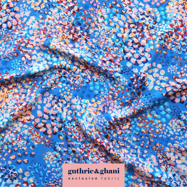 A close-up of a blue fabric with an all-over abstract floral or dot pattern in pink, orange, white, and dark blue. A pink label at the bottom reads 'guthrie&ghani exclusive fabric'.