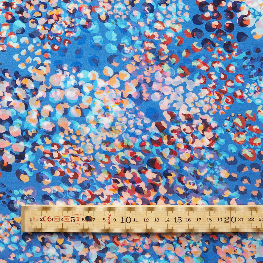 A close-up of a blue patterned fabric with a dense abstract print in pink, orange, red, white, and dark blue, with a wooden ruler along the bottom edge for scale.