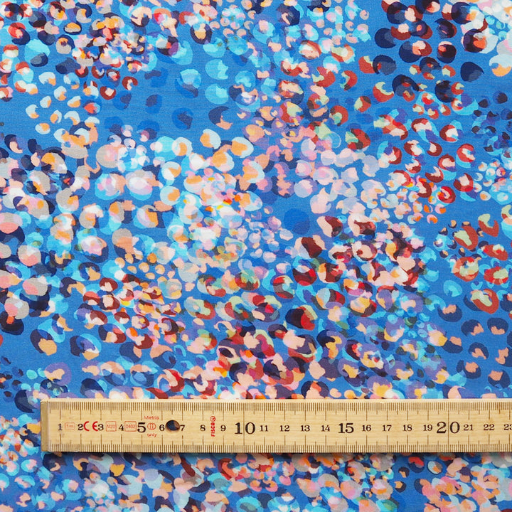 A close-up of a blue patterned fabric with a dense abstract print in pink, orange, red, white, and dark blue, with a wooden ruler along the bottom edge for scale.