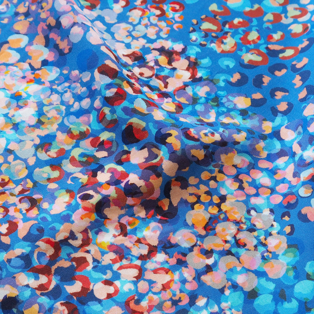 A close-up of a blue fabric with a dense, abstract leopard-like print in multiple colors including red, orange, pink, white, and dark blue.