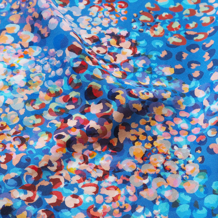 A close-up of a blue fabric with a dense, abstract leopard-like print in multiple colors including red, orange, pink, white, and dark blue.
