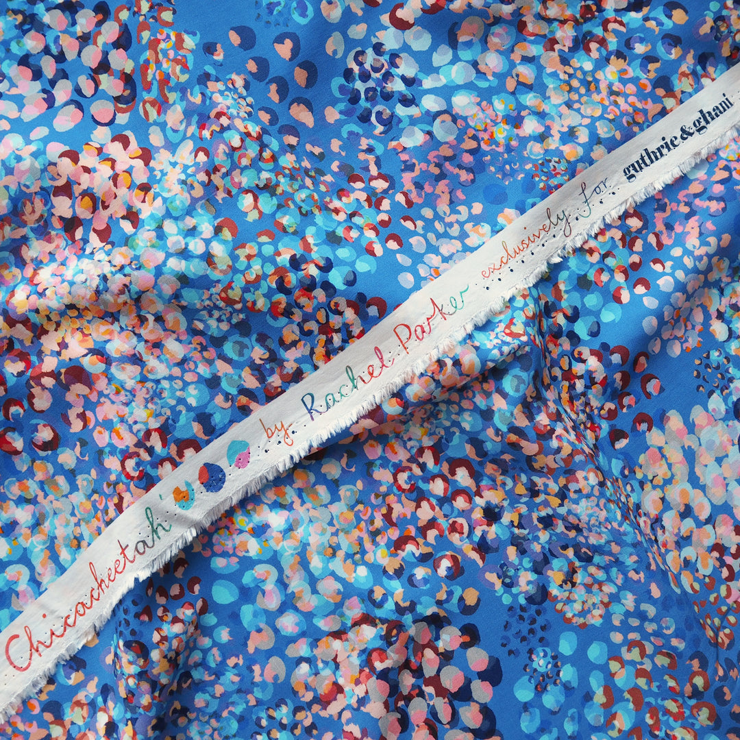 A close-up of a blue fabric with a dense multicolor floral and dot pattern, draped diagonally with a white selvedge edge showing the brand name 'Chicachetotk'.