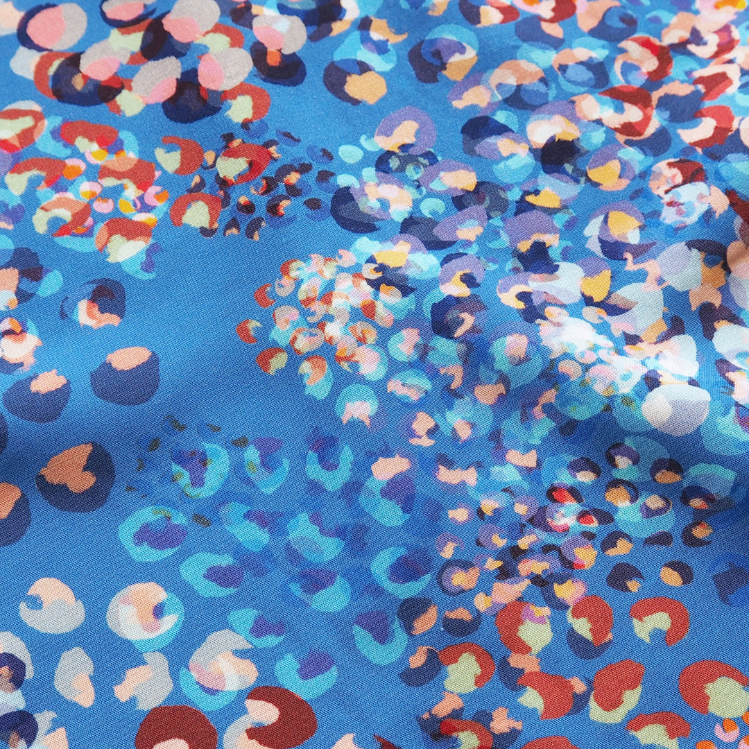A close-up of a blue fabric with a dense, abstract multicolor print featuring circles, dots, and organic shapes in red, orange, pink, purple, and white.