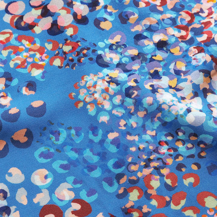 A close-up of a blue fabric with a dense, abstract multicolor print featuring circles, dots, and organic shapes in red, orange, pink, purple, and white.