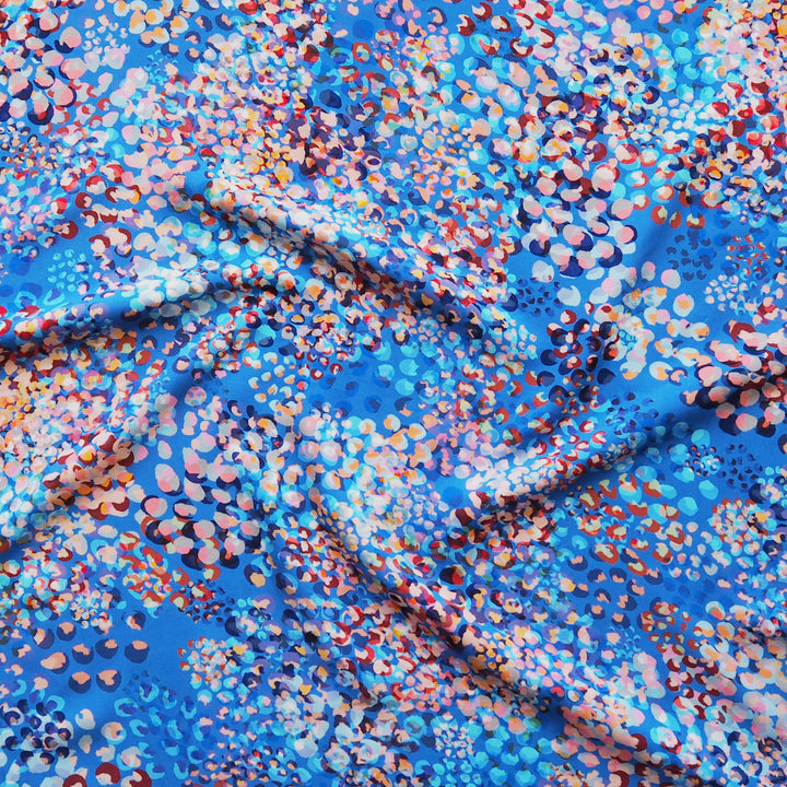 A close-up of a draped blue fabric with an all-over abstract floral or dot pattern in shades of pink, orange, red, white, and light blue.