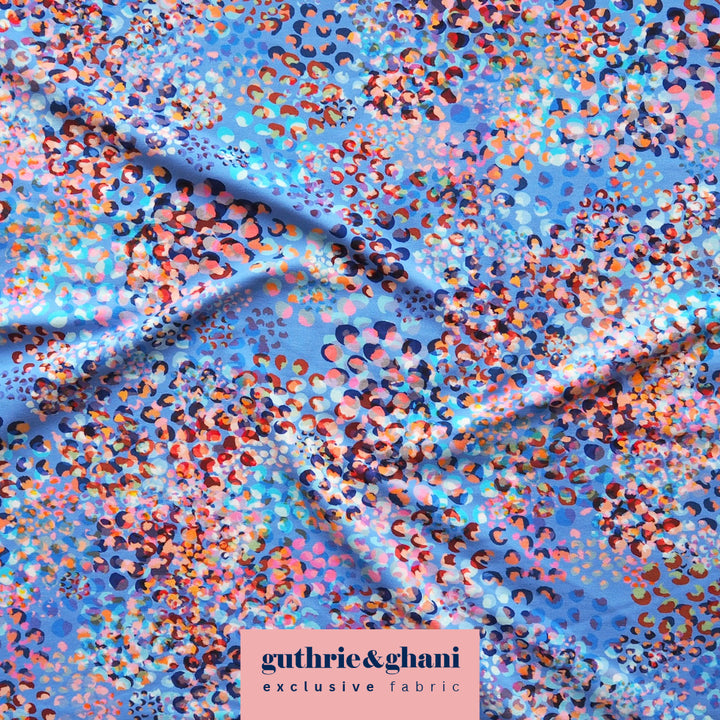 A close-up of draped fabric with a dense abstract print in light blue, pink, orange, purple, and black, with a pink label reading 'guthrie&ghani exclusive fabric' at the bottom.