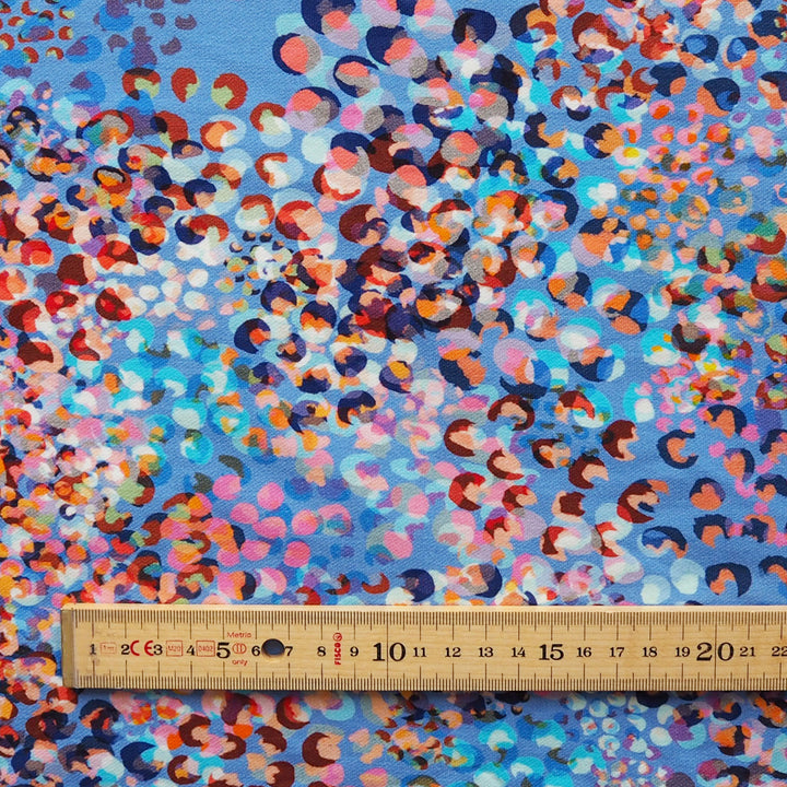 A close-up of patterned fabric with a blue background covered in dense multicolored circular spots, with a wooden ruler along the bottom edge for scale.
