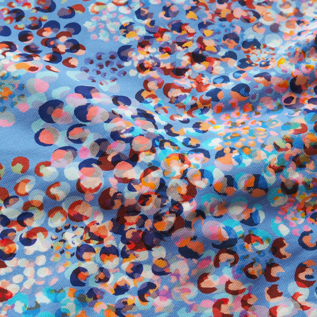 A close-up of a fabric with a light blue base and an abstract, multicolored print of overlapping circles and dots in orange, pink, red, navy, and white.