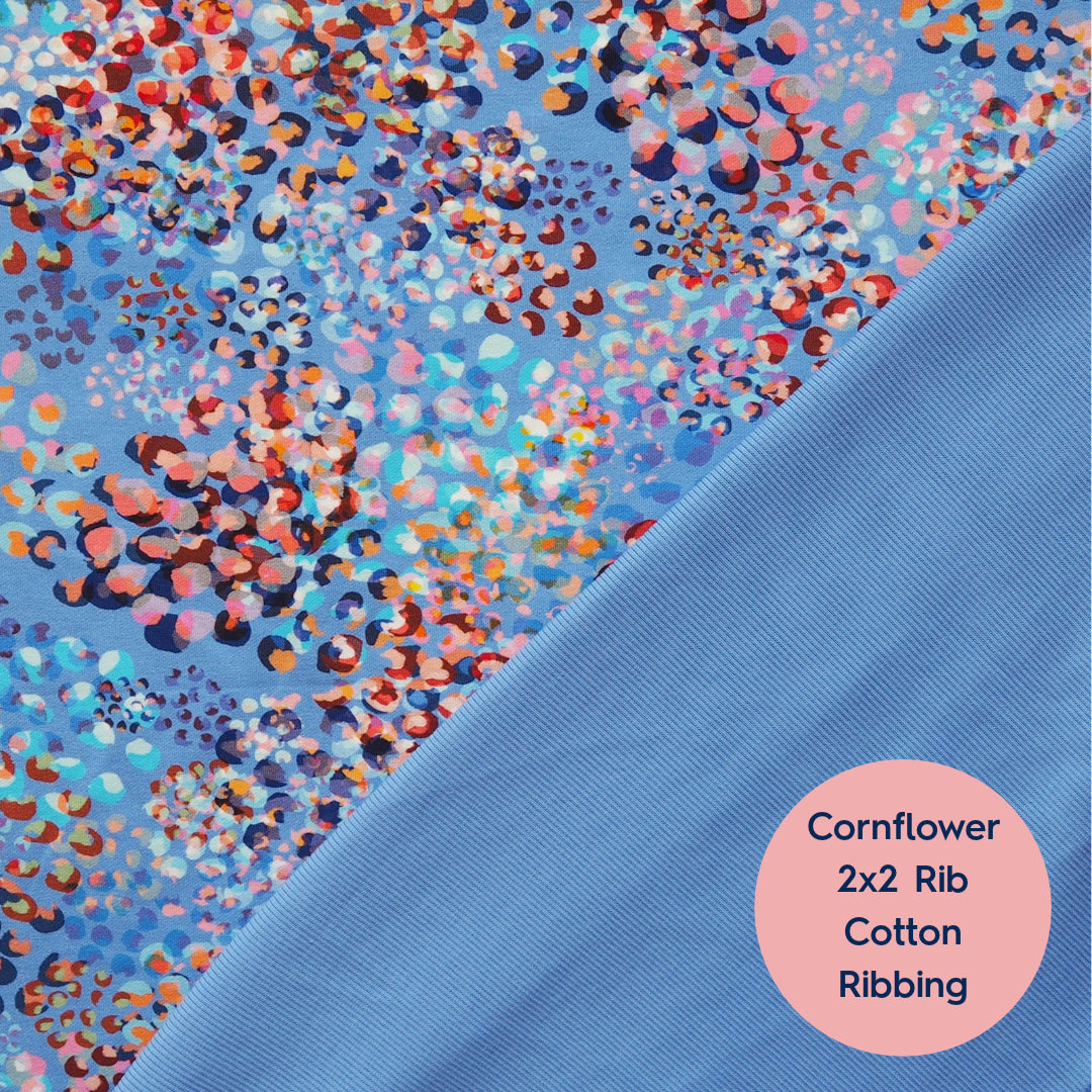 A close-up fabric sample labeled 'Cornflower 2x2 Rib Cotton Ribbing' shows a light blue ribbed section diagonally split from a larger floral print section. The print has a cornflower-like multicolor dot pattern on a light blue background.
