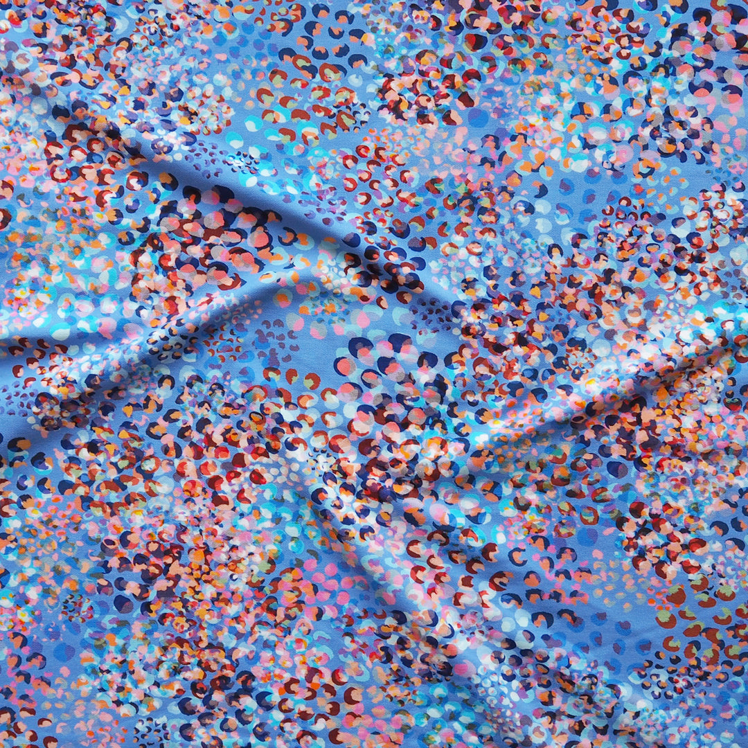 A close-up of a draped fabric with a dense, multicolored leopard-like spot pattern on a light blue base.