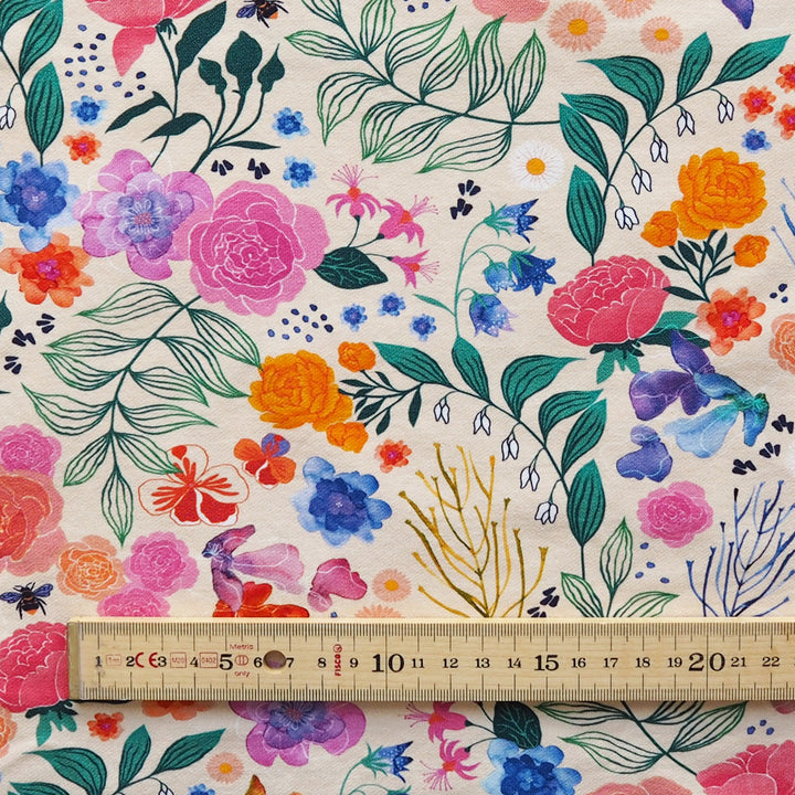 A close-up of a floral fabric pattern on a cream background, featuring watercolor-style pink, orange, blue, purple, green, and gold botanical motifs. A wooden ruler is placed along the bottom edge to show scale.