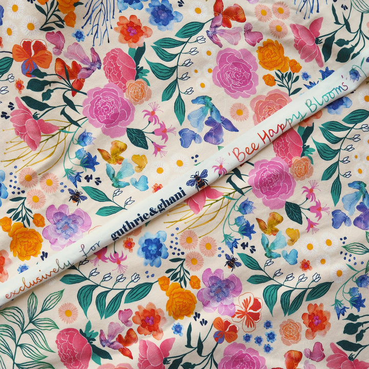 A close-up of a floral fabric with a colorful watercolor print on a cream background, featuring pink, orange, blue, purple, and green flowers and leaves. A white fabric marker labeled 'suthrie&dhani' and 'Bee Happy Blossom' is placed diagonally across the fabric.