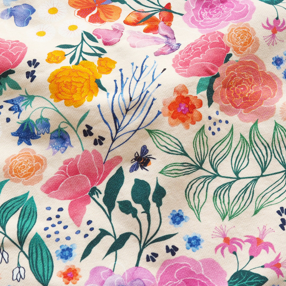 A close-up of a fabric or wallpaper pattern featuring a colorful watercolor floral design on a cream background. The print includes pink, orange, yellow, blue, and green botanical elements such as roses, leaves, flowers, and a small bee.