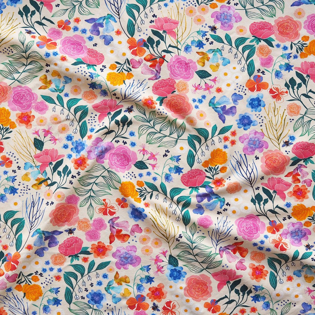 A close-up of a draped fabric featuring a vibrant, multicolor floral print on a warm cream background. The pattern includes pink, orange, blue, purple, and green watercolor-style flowers and leaves.