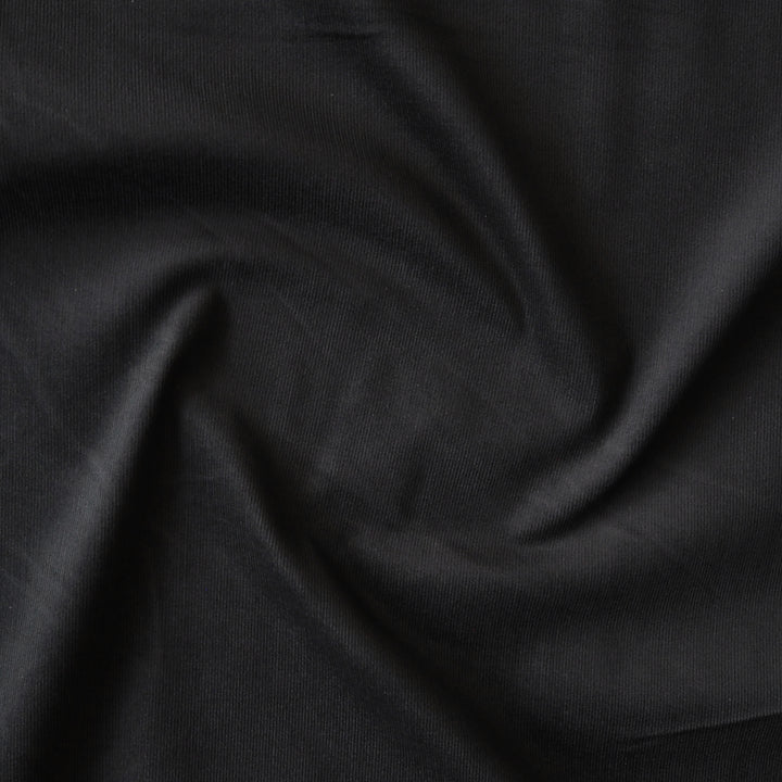 Black stretch cotton needlecord fabric, soft fine cord texture.