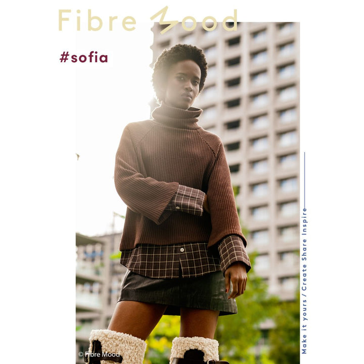 Person wearing a brown ribbed sweater with plaid cuffs and hem, standing in an urban setting.