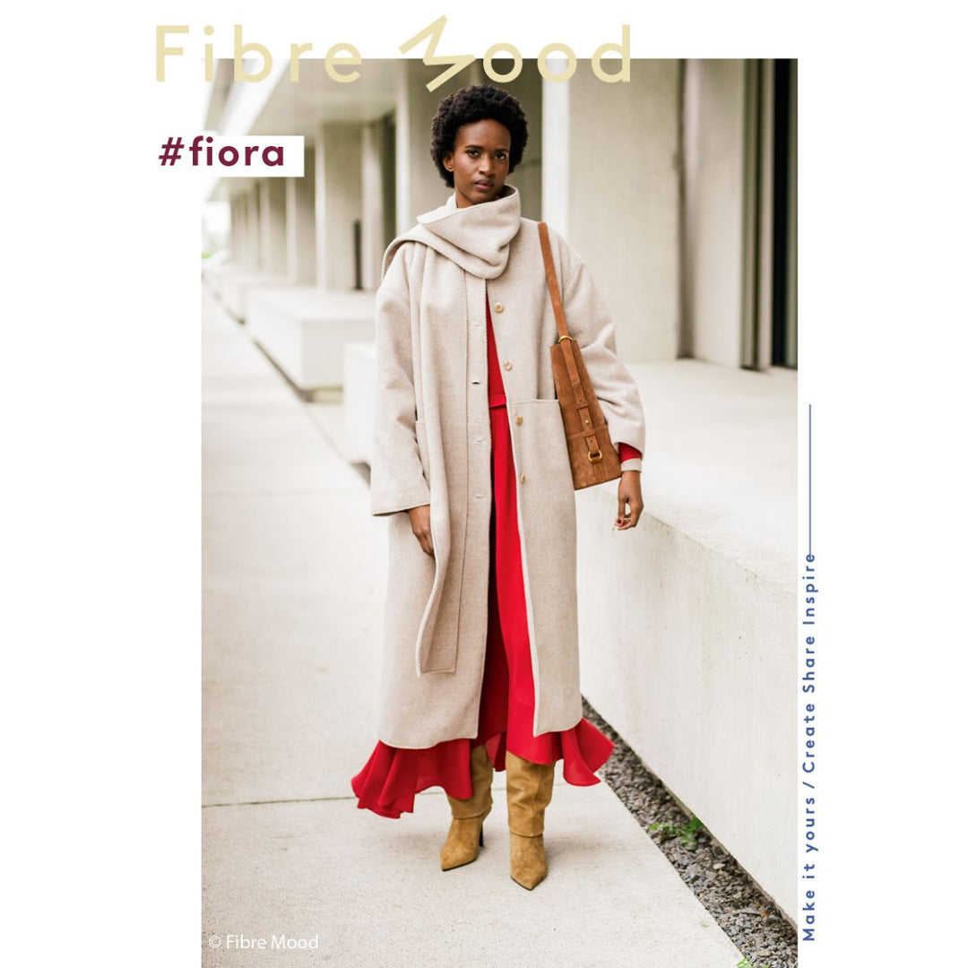Woman wearing a long beige coat over a red dress with a brown bag, standing on a sidewalk.