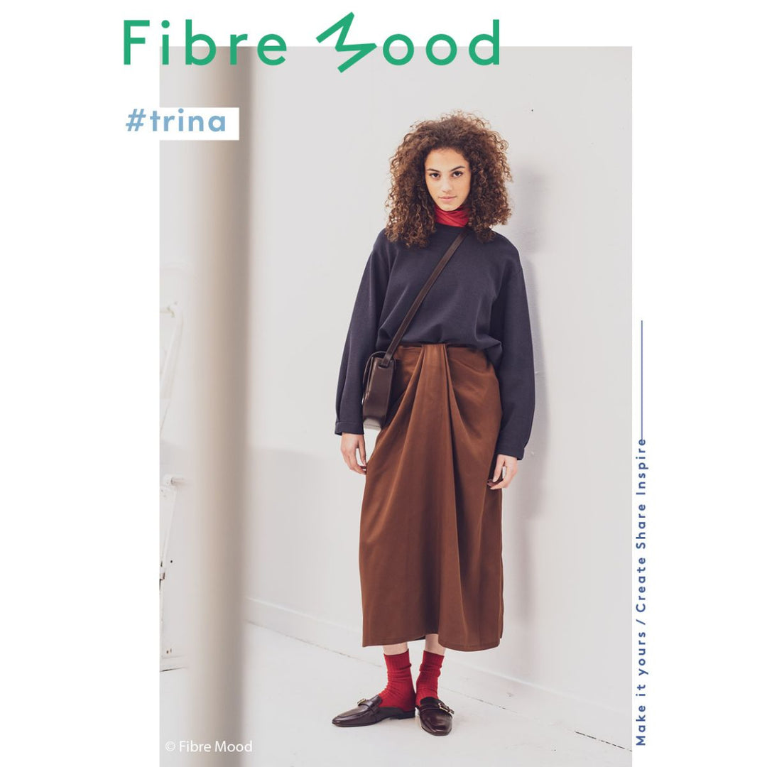 Woman wearing a dark sweater and brown skirt with a crossbody bag, standing against a white wall with 'Fibre Mood' branding.