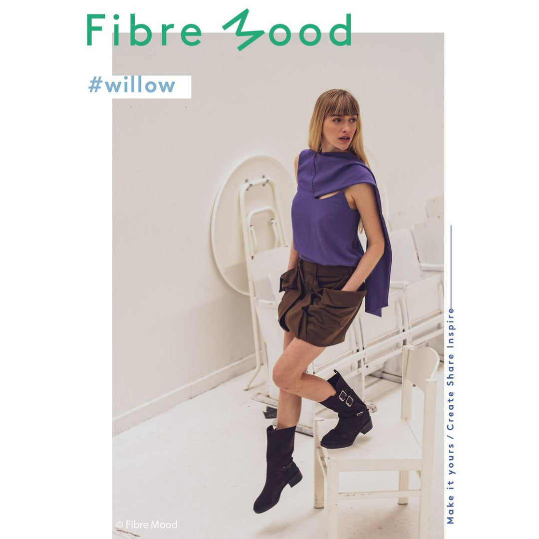 Woman in a purple top and brown skirt with black boots standing in a minimalistic room, wearing a scarf. 'Fibre Mood' brand visible.