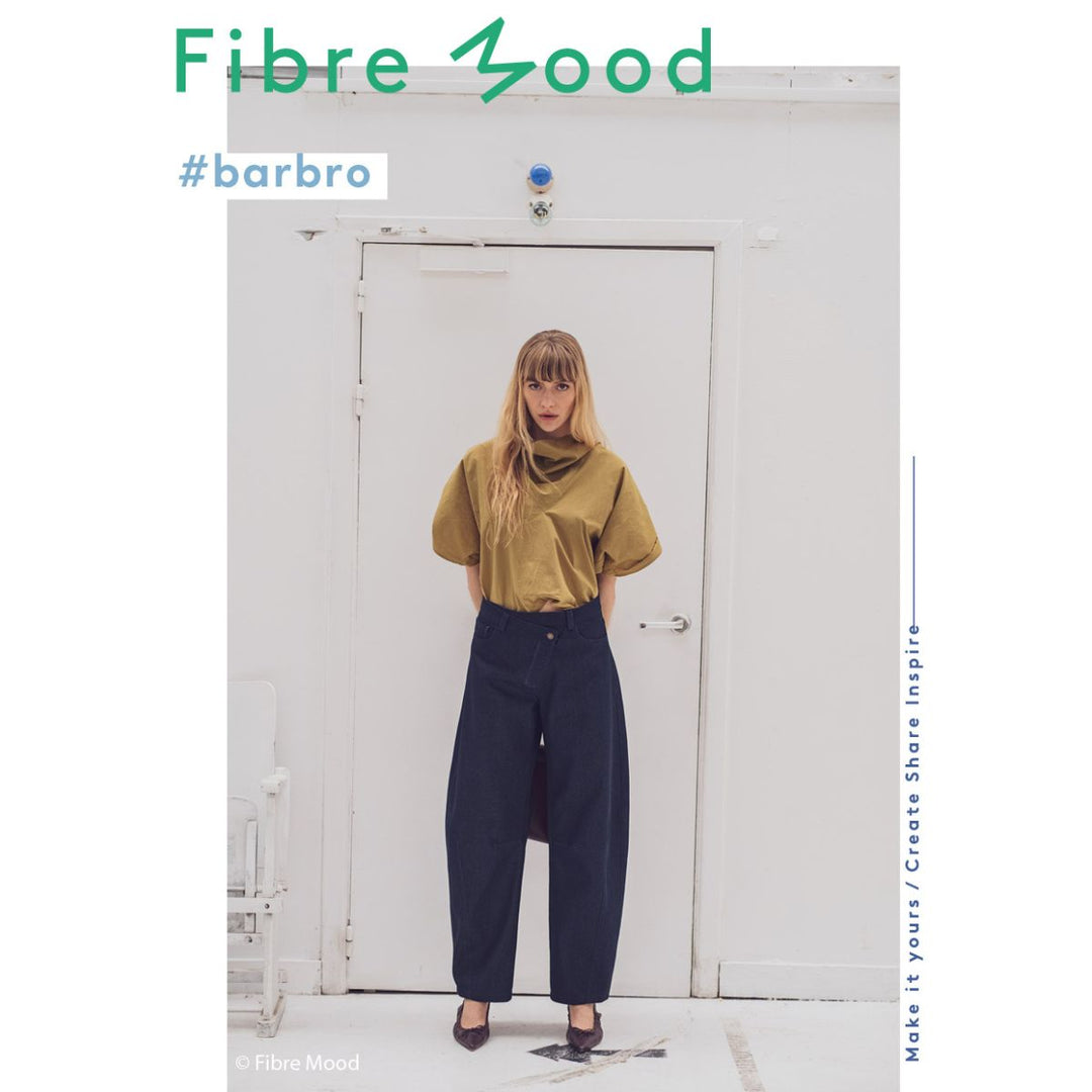 Person wearing a mustard yellow top and dark blue pants standing in front of a white door with 'Fibre Mood' branding.