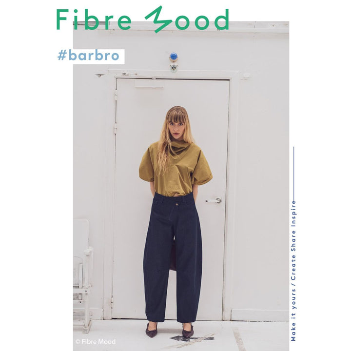 Person wearing a mustard yellow top and dark blue pants standing in front of a white door with 'Fibre Mood' branding.