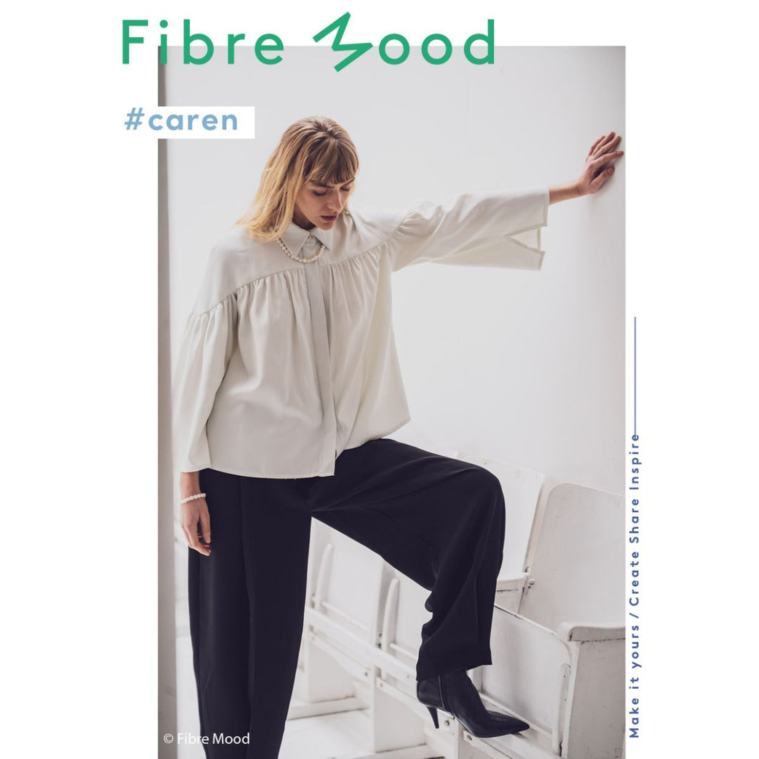 Woman wearing a light blouse and dark pants, leaning against a white wall with 'Fibre Mood' branding.