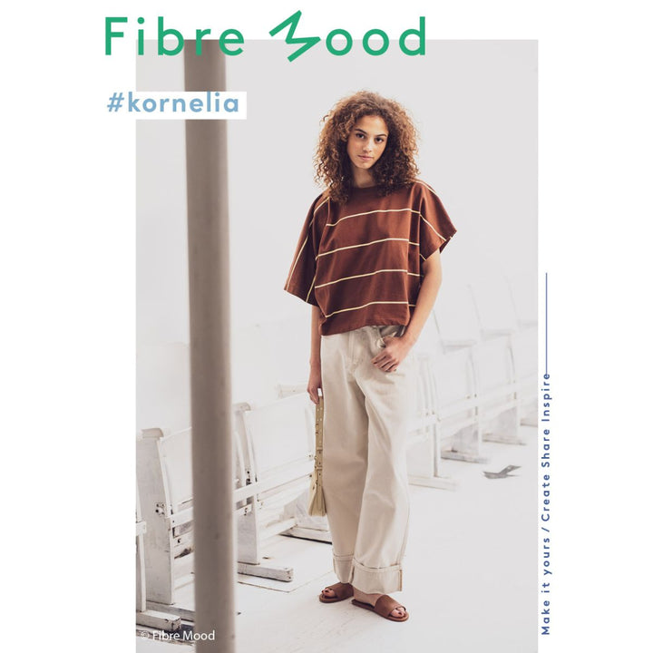 Woman wearing a brown striped poncho and white pants in a minimalistic room with 'Fibre Mood' branding.