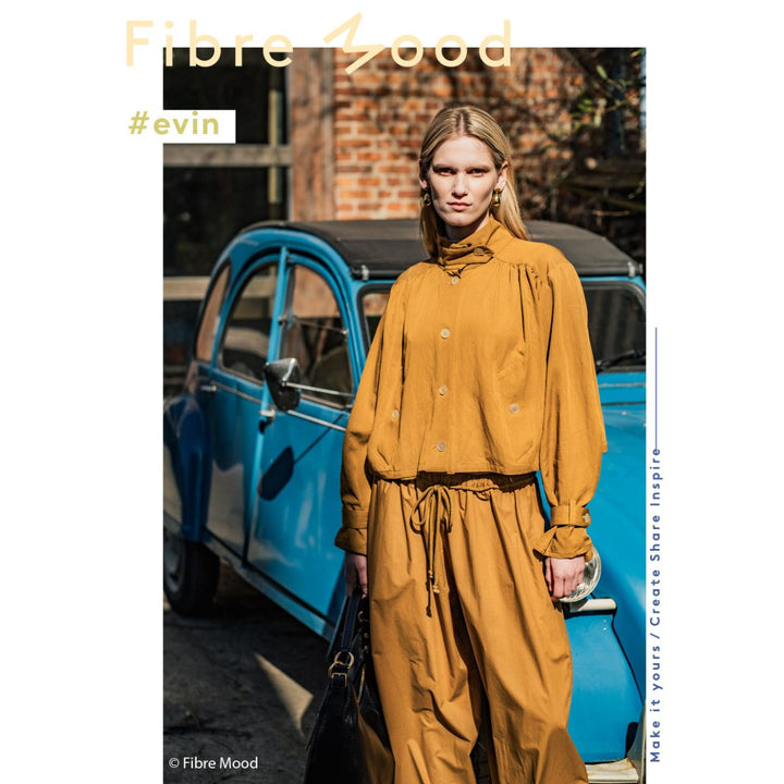 Woman in mustard yellow outfit standing next to a blue car with 'Fibre Mood' branding.