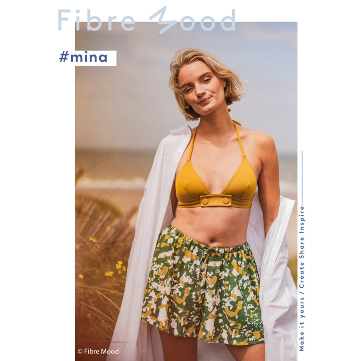 Woman wearing a yellow bikini top and green floral shorts with a white cover-up on a beach.